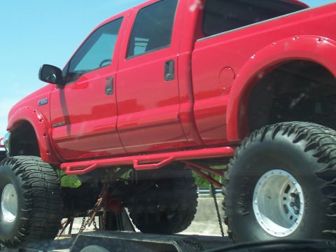 2004 Aston Martin Virage - Lifted red pickup truck with large off-road tires is being transported on a trailer.