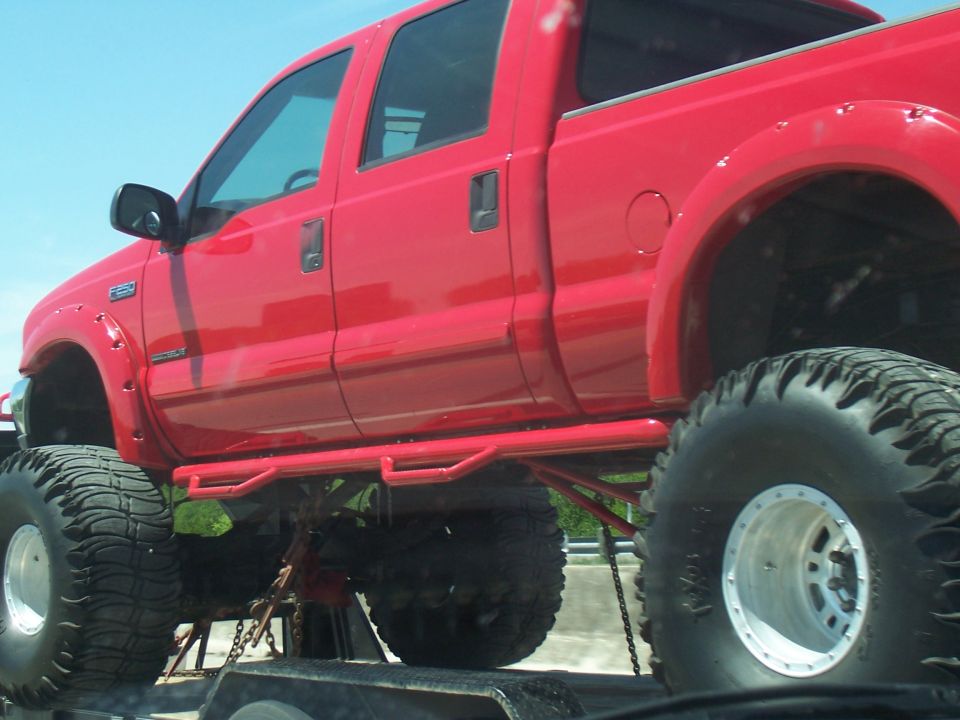 Red pickup truck with oversized tires and custom side steps, loaded on a trailer