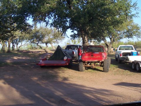2004 Aston Martin Virage - Two pickup trucks and a trailer are parked under trees in a dirt lot.
