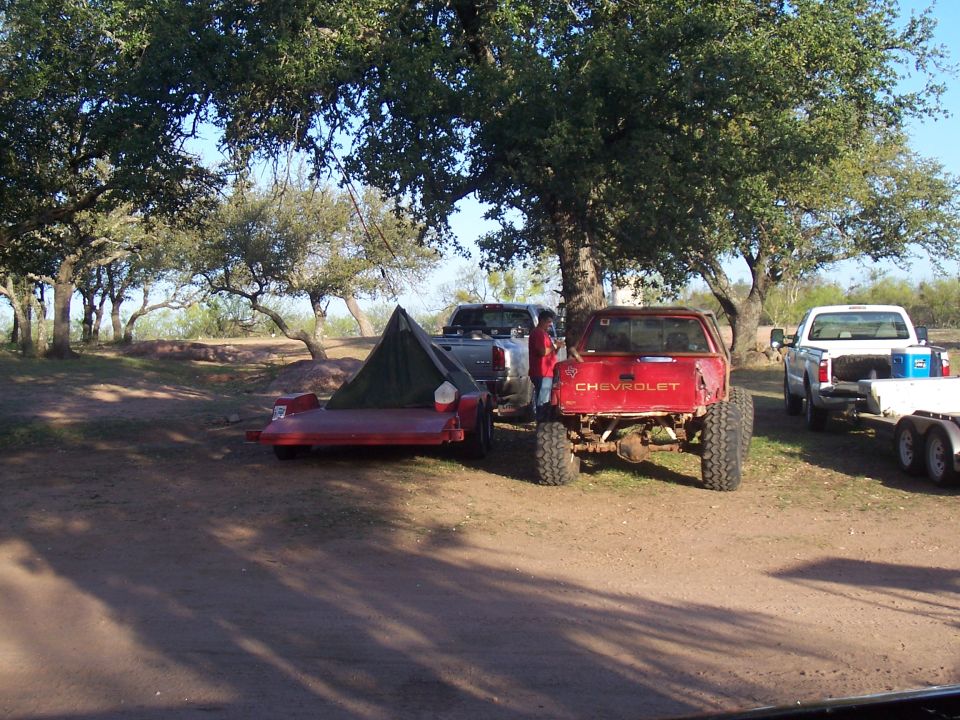 Red Chevrolet pickup truck with large tires, rear view. A green tent is on a red trailer next to it. A silver pickup truck is also visible