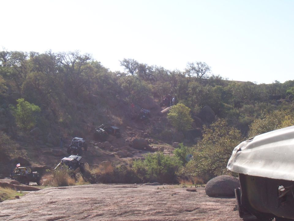 Several off-road vehicles navigate a steep, rocky incline with sparse trees and large boulders