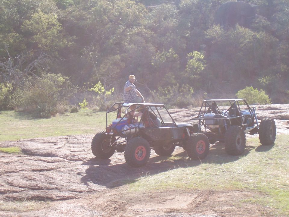 Two off-road buggies parked on rocky ground with trees in the background. One buggy has a person sitting on top