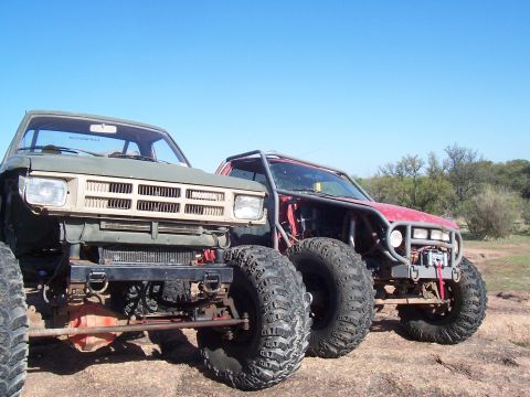 2004 Aston Martin Virage - Two heavily modified off-road trucks sit parked on a rocky terrain under a clear blue sky.