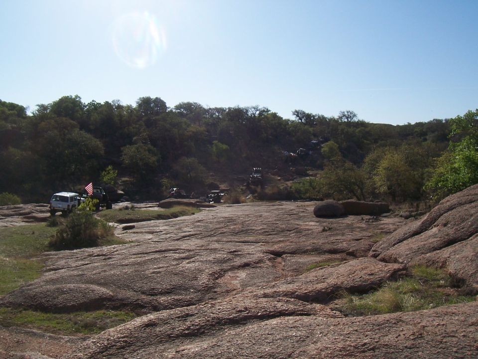 Several off-road vehicles parked on a rocky, uneven landscape with trees and a clear blue sky