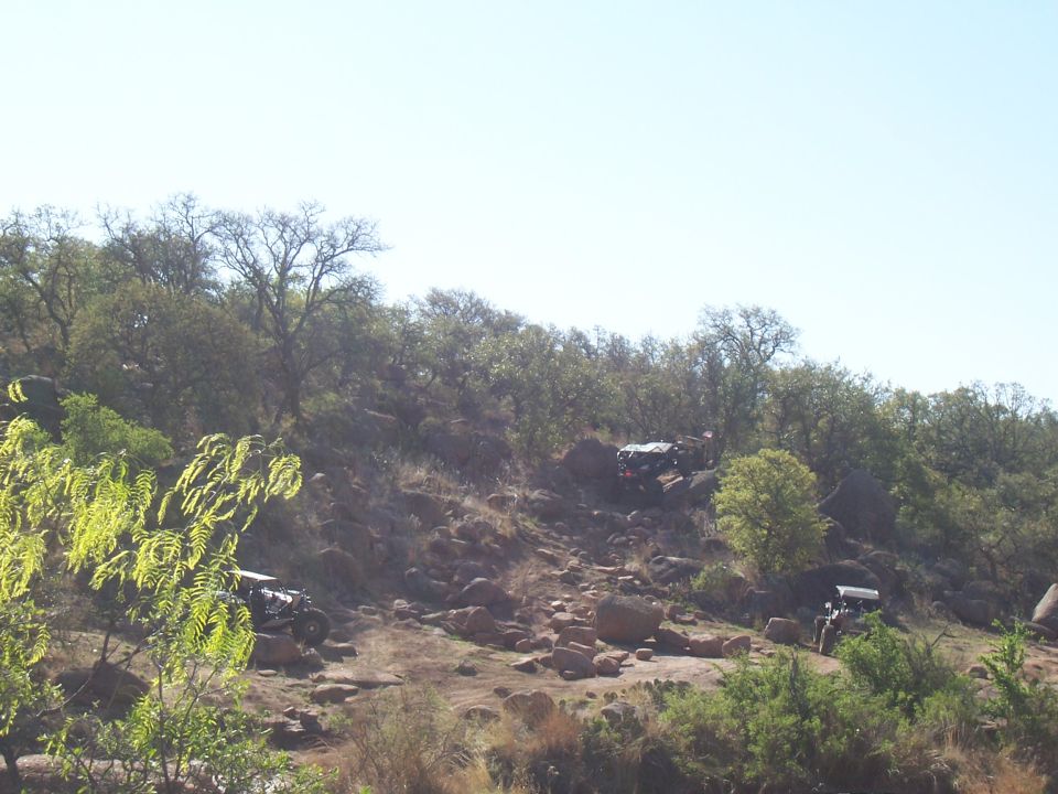 Three off-road vehicles navigate a steep, rocky hill with sparse trees and clear sky