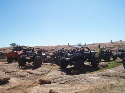 2004 Aston Martin Virage - A collection of custom off-road vehicles are parked on a rocky terrain, with several individuals visible in the background.