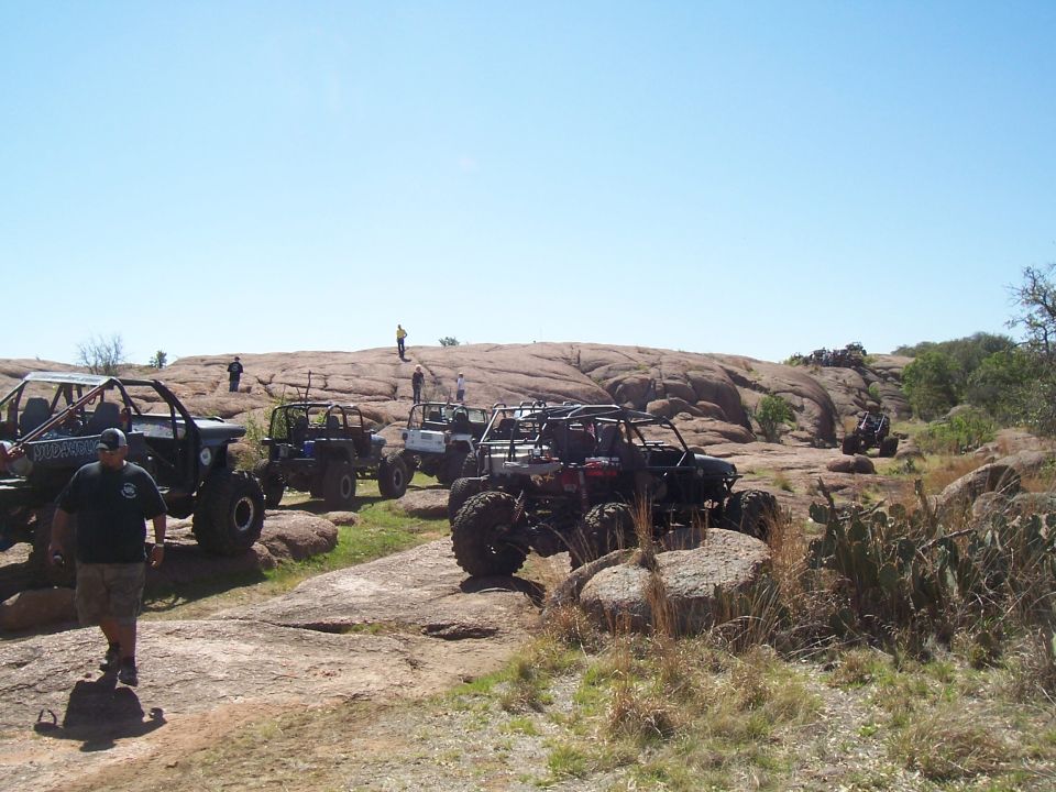 Several off-road buggies are parked and driving on a large, rocky hill with a clear blue sky above