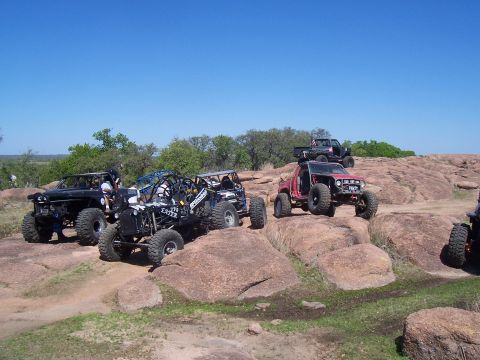 2004 Aston Martin Virage - Multiple modified off-road vehicles navigate a challenging rocky landscape on a sunny day.
