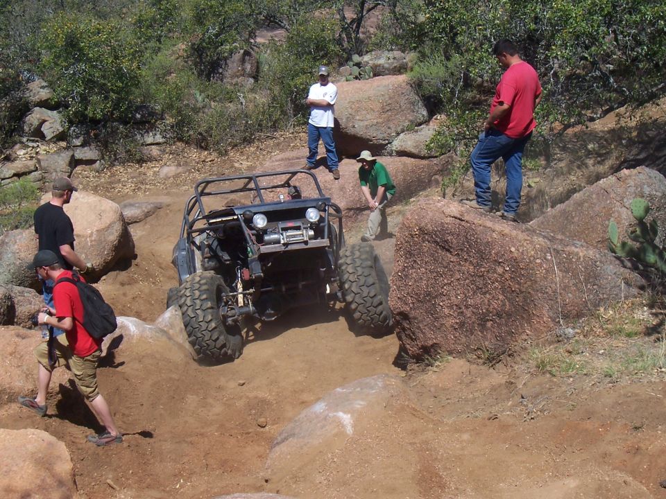 Off-road buggy with a roll cage and large tires climbing a rocky hill. Several people are watching