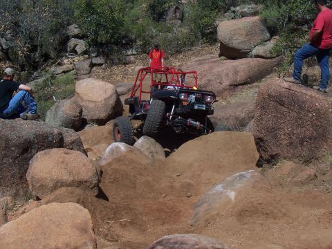 2004 Aston Martin Virage - An off-road buggy with a prominent red roll cage navigates a challenging rocky trail, with spectators observing the action.