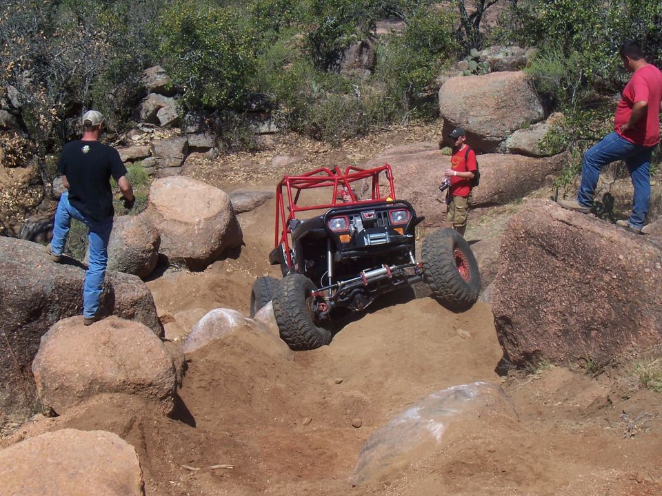 Off-road buggy with red roll cage climbing over rocks, two people watching