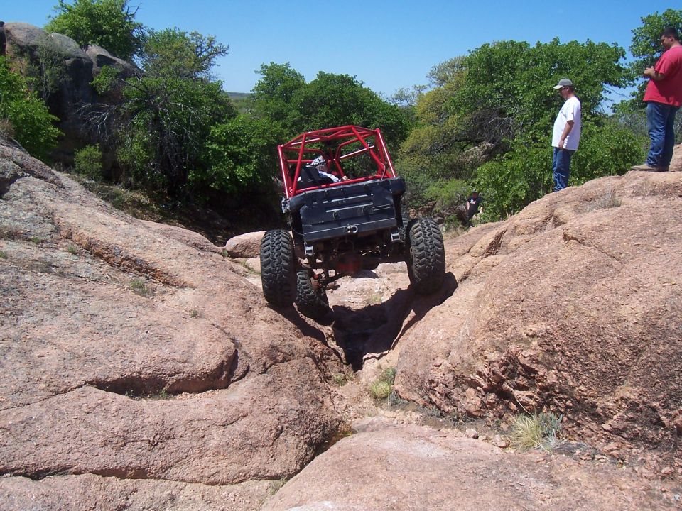 Off-road vehicle with a red roll cage climbing over large rocks on a sunny day. Two people watch from a rocky outcrop