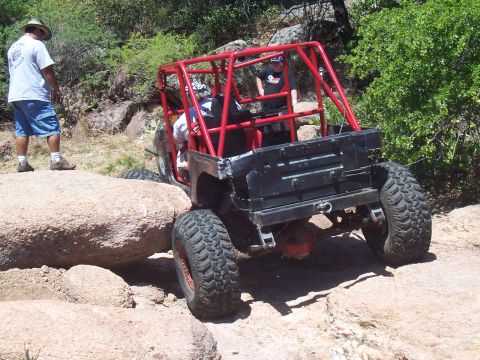 2004 Aston Martin Virage - An off-road vehicle with a prominent red roll cage maneuvers over large rocks on a trail.