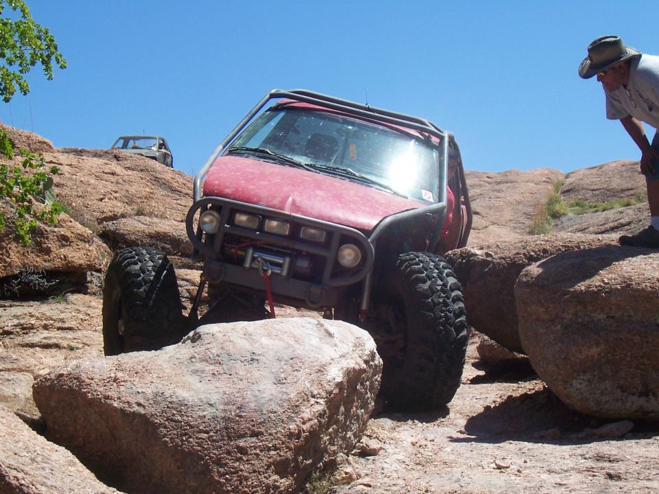 Red off-road vehicle with roll cage navigating rocky terrain; another vehicle visible on ridge above
