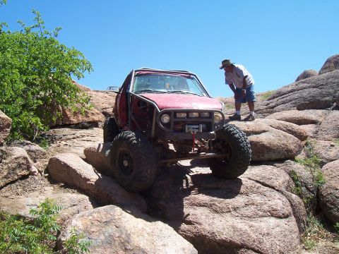 2004 Aston Martin Virage - A man watches a red off-road vehicle with a roll cage as it maneuvers over large rocks on a sunny day.