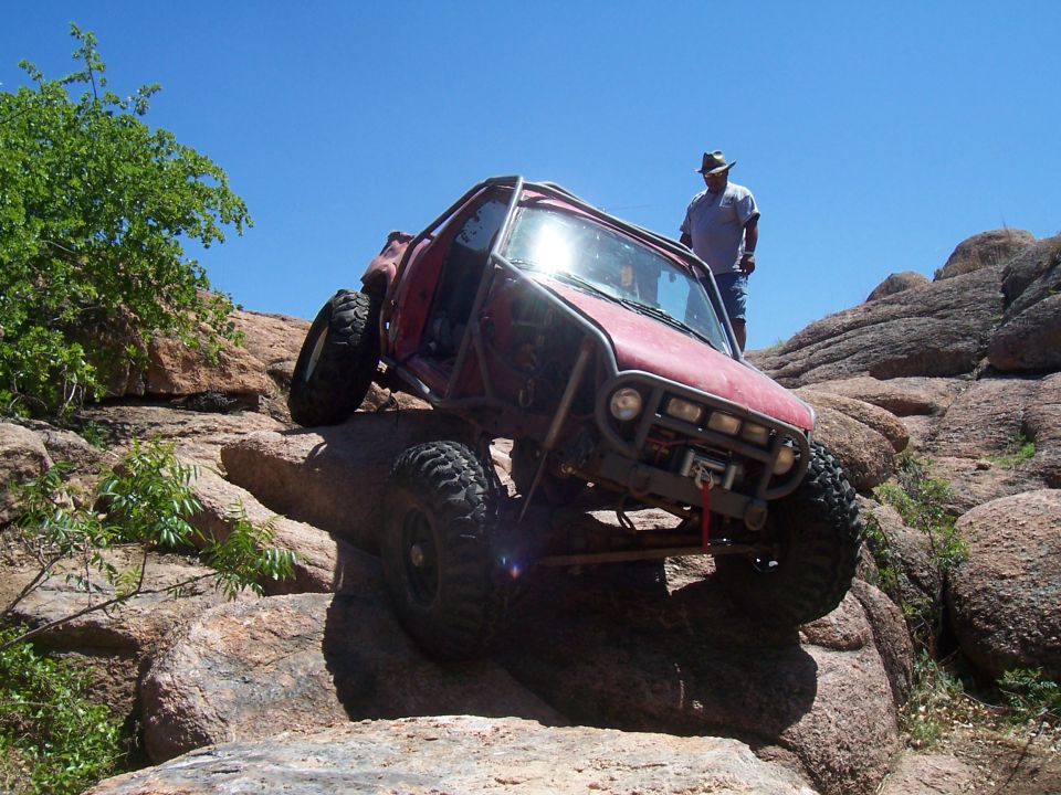 Red off-road buggy with a roll cage is climbing over large rocks. A man stands on a rock behind it