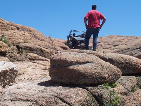 2004 Aston Martin Virage - A person stands on a rock overlooking an off-road buggy navigating a steep, rocky incline under a clear blue sky.