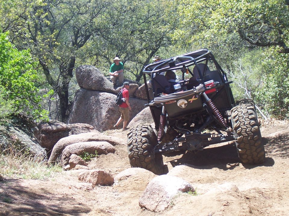 Black off-road buggy with a roll cage navigates a rocky, dirt trail with two people watching