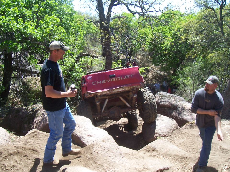 Man watches red Chevrolet pickup truck navigate rocky terrain. Another man stands nearby