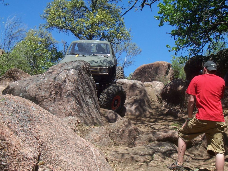 Green off-road vehicle navigating a rocky trail with a person in a red shirt watching