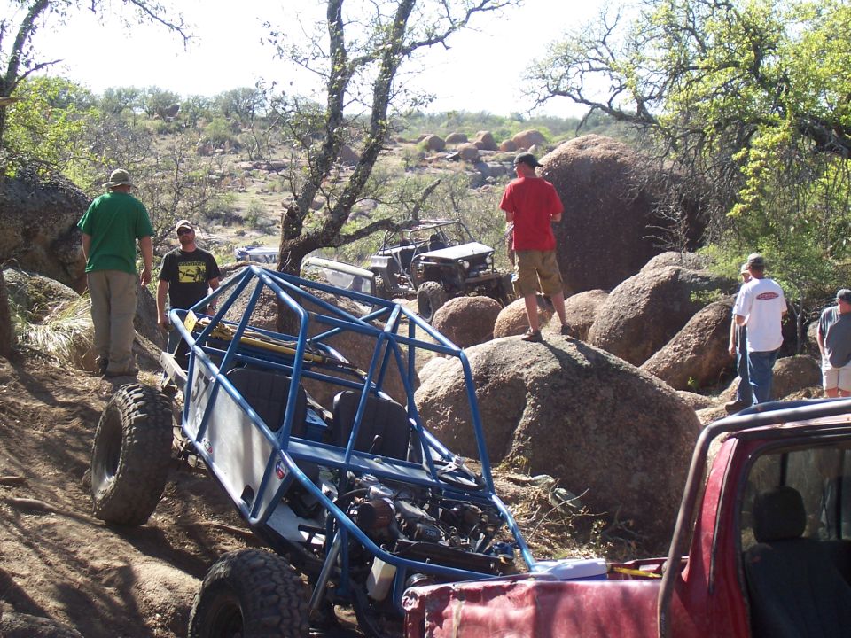Off-road buggy with a roll cage on a dirt trail, other vehicles and people in background