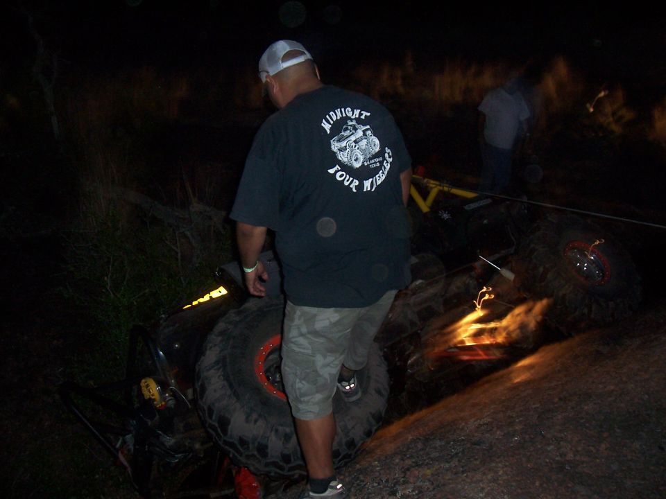 Overturned off-road vehicle with large tires and orange accents, at night, with people nearby