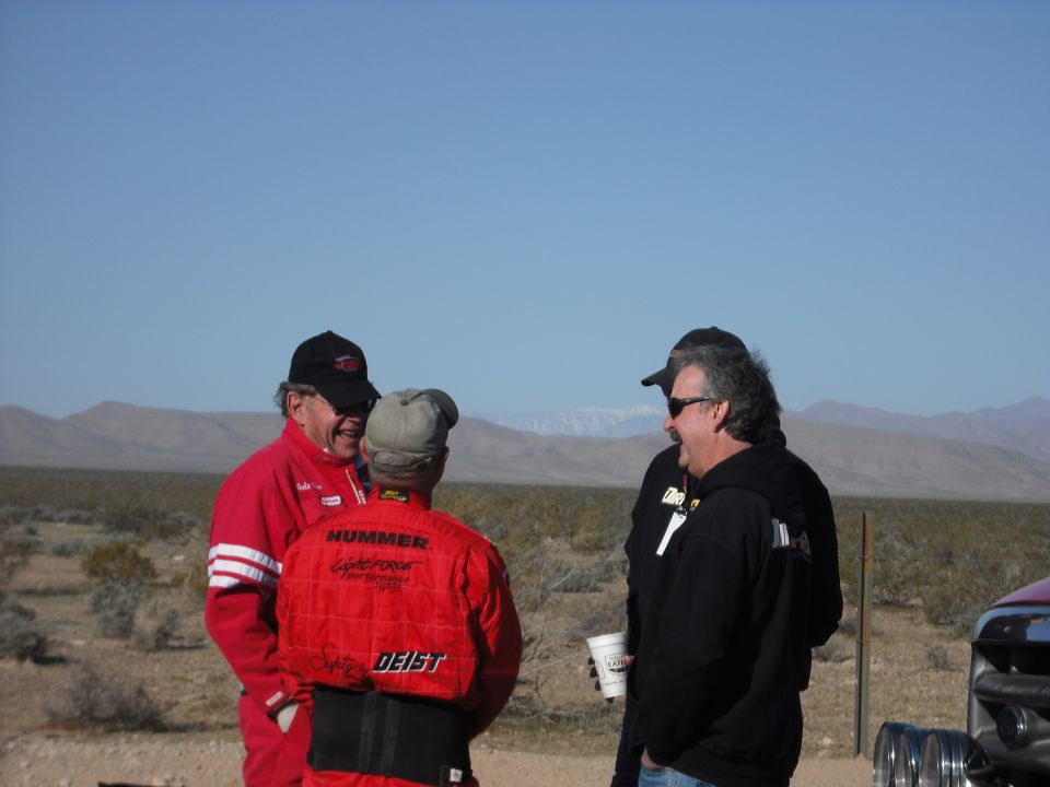 Three men in racing attire talking in a desert with mountains in the background.