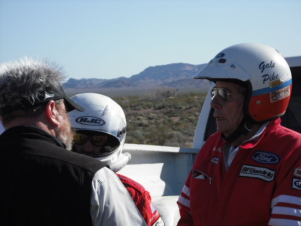 Two men in racing gear and helmets stand in the bed of a white pickup truck.