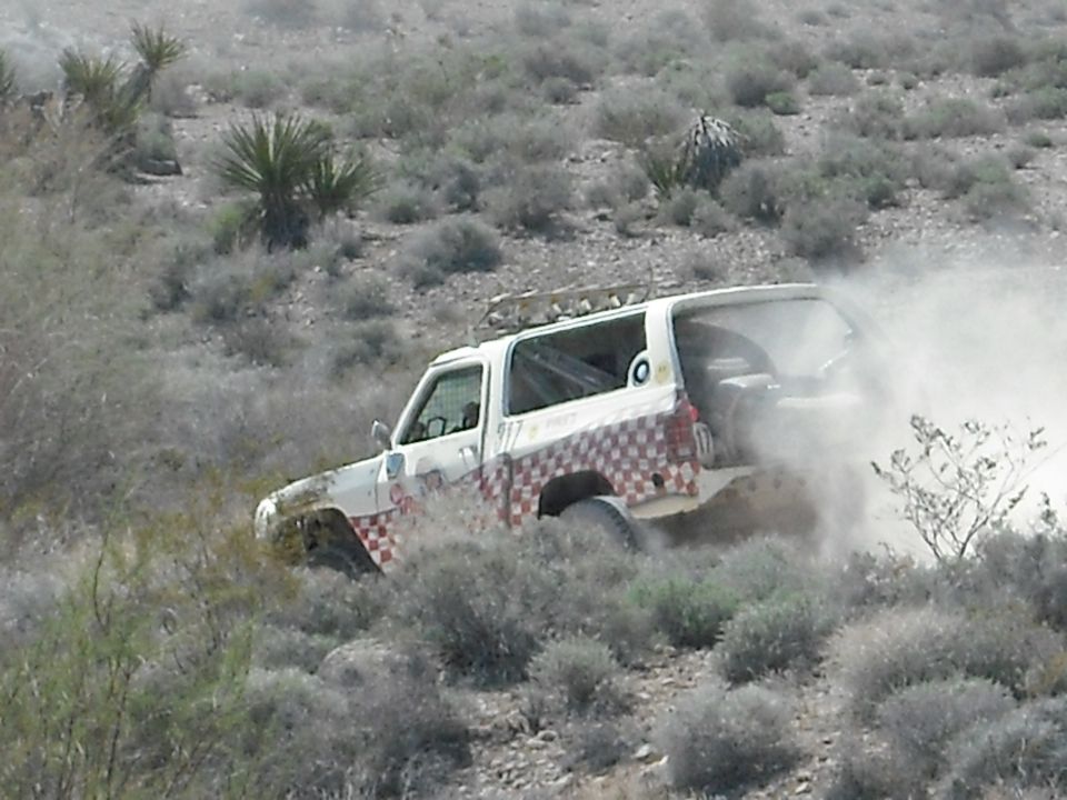 White off-road vehicle with checkered pattern driving through dusty brush.