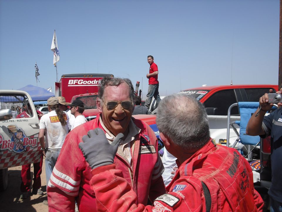 Two men in red racing suits, one giving a thumbs up, in front of a red truck.