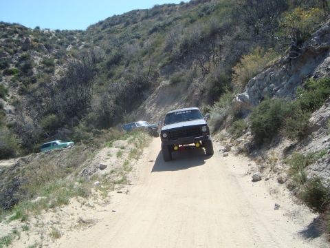 1986 Ford Bronco - A gray 1986 Ford Bronco is driving on a dirt road in a hilly, arid landscape. Another blue SUV is visible further up the road.