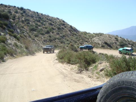 1986 Ford Bronco - A dark-colored Ford Bronco is driving on a dirt road in a hilly, arid landscape. Another green SUV is visible further down the road.
