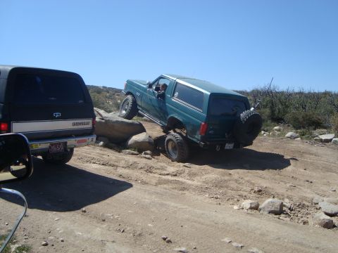 1986 Ford Bronco - A teal Ford Bronco is navigating a rocky, off-road trail, with its front passenger side tire climbing over a large boulder. Another black S…