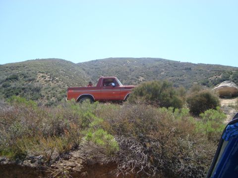 1986 Ford Bronco - A red Ford pickup truck is parked on a dirt road surrounded by dry brush and hills under a clear blue sky. Two people are visible in the ca…