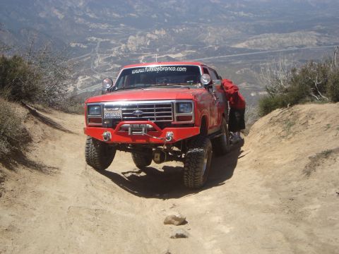 1986 Ford Bronco - A red 1980s Ford Bronco is parked on a dirt trail in a mountainous, arid landscape. The vehicle has a custom front bumper with a winch and…