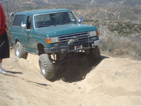 1986 Ford Bronco - A teal 1986 Ford Bronco is shown off-roading on a dirt trail, with its front tires angled up a sandy incline. The vehicle has large off-roa…