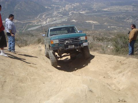 1986 Ford Bronco - A teal Ford Bronco is driving off-road on a dirt trail with two people standing on the side of the trail watching.