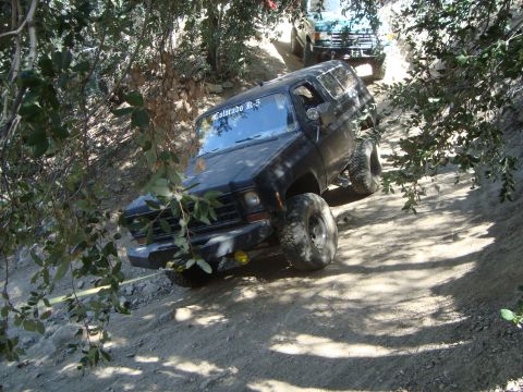 1986 Ford Bronco - A black 1980s SUV is driving up a steep, dirt trail surrounded by trees. The SUV has large off-road tires and a yellow tow strap attached t…
