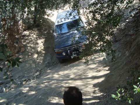 1986 Ford Bronco - A dark blue Ford Bronco is driving up a steep, dirt trail surrounded by trees. The vehicle has a winch mounted on the front bumper and text…
