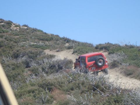1986 Ford Bronco - A red 1986 Ford Bronco is driving up a dirt path on a hillside covered in dry brush and sparse vegetation under a clear blue sky.