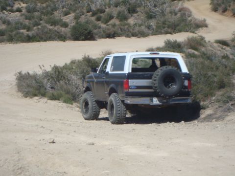 1986 Ford Bronco - The rear driver's side of a dark-colored Ford Bronco with a white roof is parked on a dirt road. A spare tire is mounted on the rear door,…