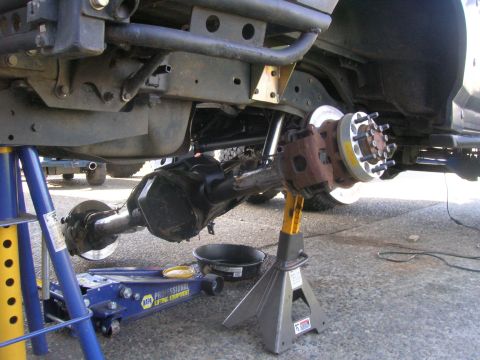 1996 Ford Bronco - The underside of a vehicle is shown, supported by jack stands and a floor jack. A rear axle assembly with brake components and a wheel hub…