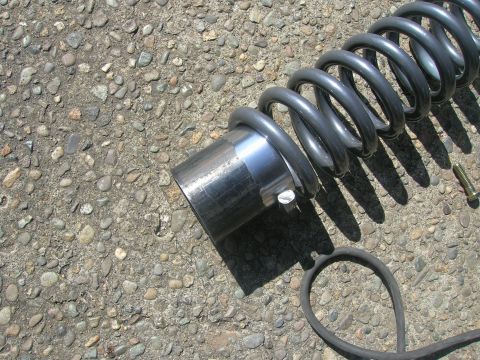 1996 Ford Bronco - A close-up view of a heavy-duty coil spring with a metal sleeve on a gravelly surface. A bolt and a black rubber hose are also visible in t…