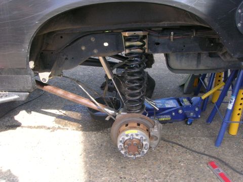 1996 Ford Bronco - Front suspension of a jacked-up SUV with the wheel removed, showing a black coil spring over a shock, radius arm, brake caliper and rusty r…