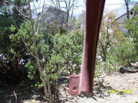 1972 Ford Ranchero - A close-up of a rusty metal bracket in the foreground obscures a view of green bushes and trees, with a house visible in the background.