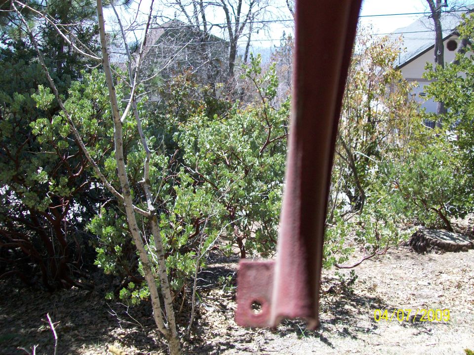 Rusty metal bracket in foreground with green bushes and trees behind it.