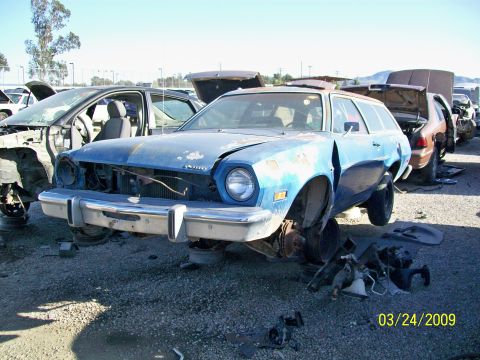 1972 Ford Ranchero - A light blue station wagon is parked in a junkyard with its hood open and front end damaged. Other vehicles and car parts are visible in th…