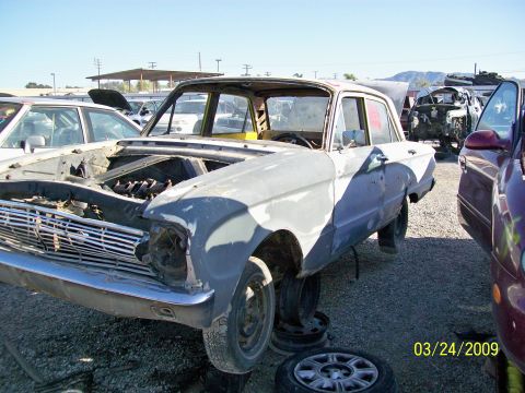 1972 Ford Ranchero - A light gray, two-door sedan is parked at a junkyard with its hood open, revealing the engine bay. The car is missing its front headlights…