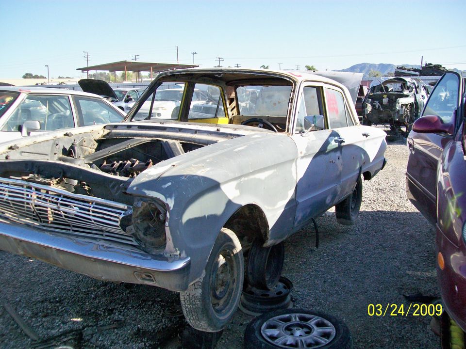 Front 3/4 view of a light gray sedan at a junkyard, missing headlights and with hood open.