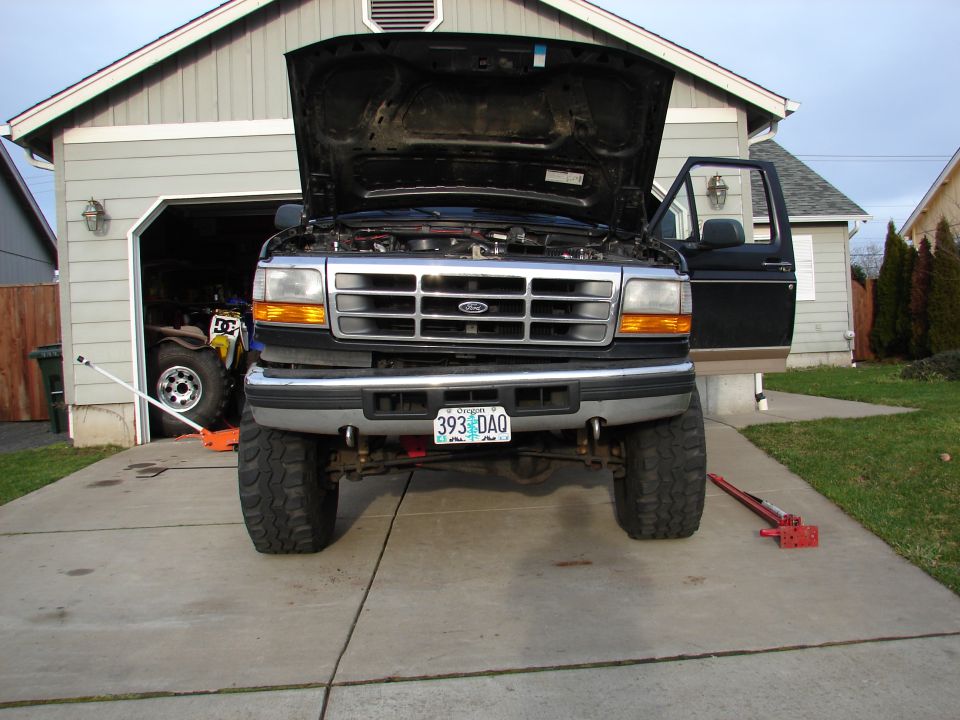 Black Ford pickup truck with open hood lifted on jack in driveway